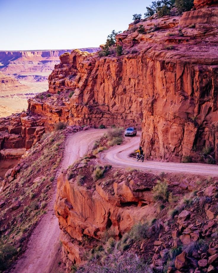 This may contain: a car driving down a dirt road next to mountains and cliffs in the desert at sunset