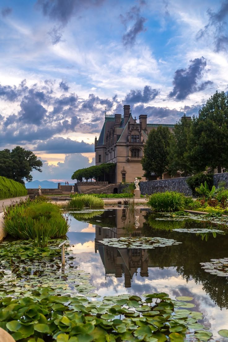 This may contain: a large house sitting on top of a lush green field next to a pond filled with water lilies