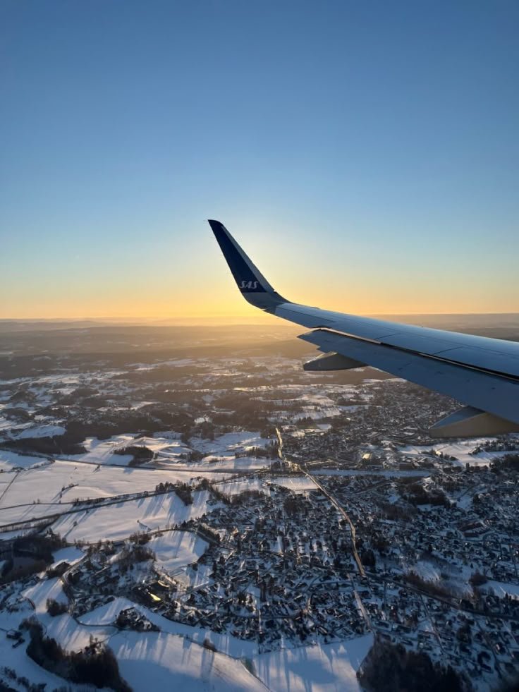This may contain: the wing of an airplane as it flies over snow - covered fields and trees at sunset