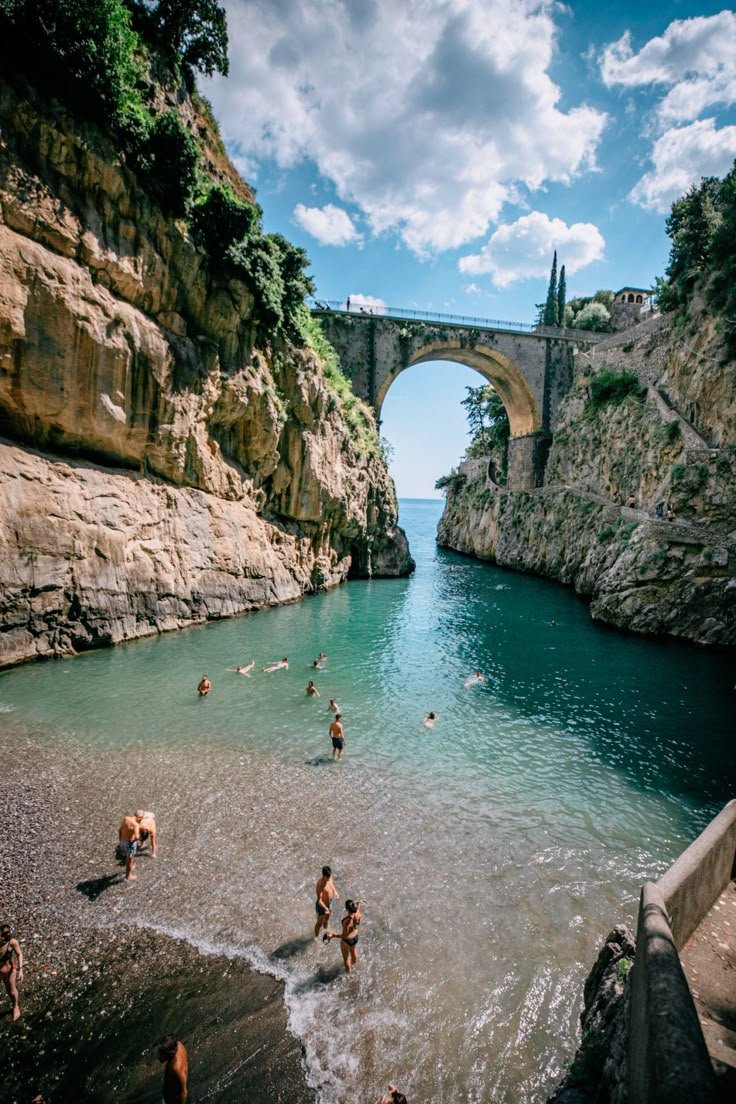 This may contain: people are playing in the water near an old stone bridge and some cliffs on either side