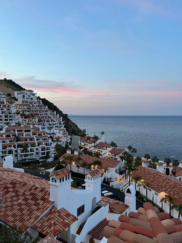 This may contain: an aerial view of houses and the ocean at dusk, with mountains in the background