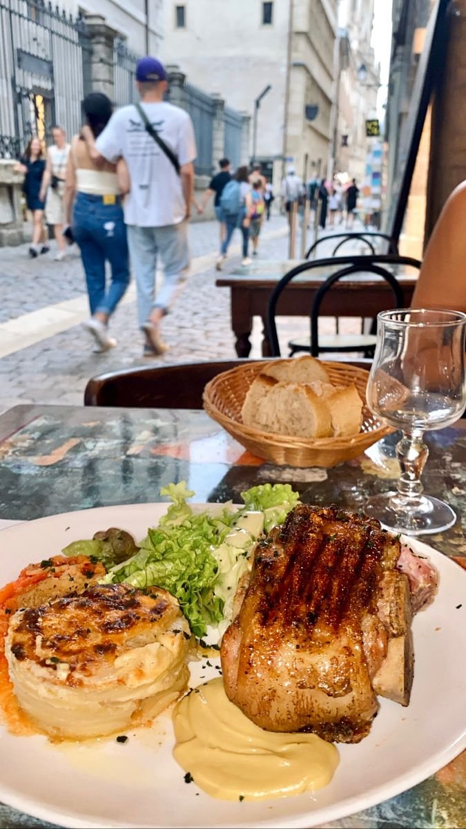 This may contain: a white plate topped with meat and vegetables next to a glass of wine on a table