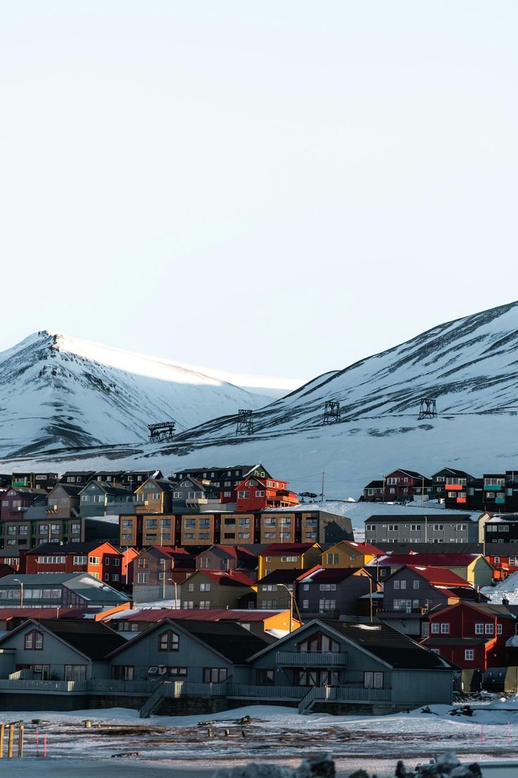 This may contain: a town with mountains in the background and snow on the ground around it, as seen from across the water