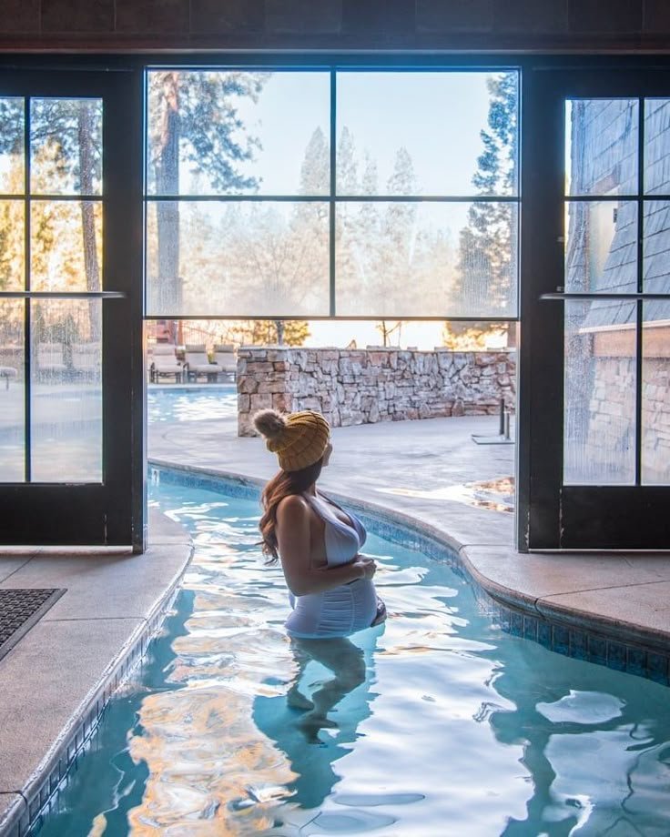 This may contain: a woman sitting on the edge of a swimming pool in front of a large window