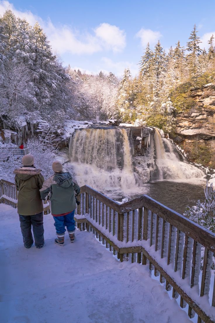 This may contain: two people standing on a bridge looking at a waterfall in the snow with trees around