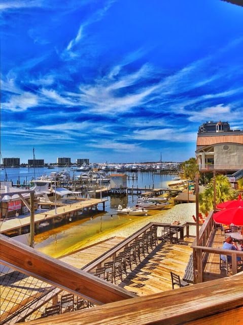 This may contain: an outdoor deck with tables and umbrellas next to the water in front of boats