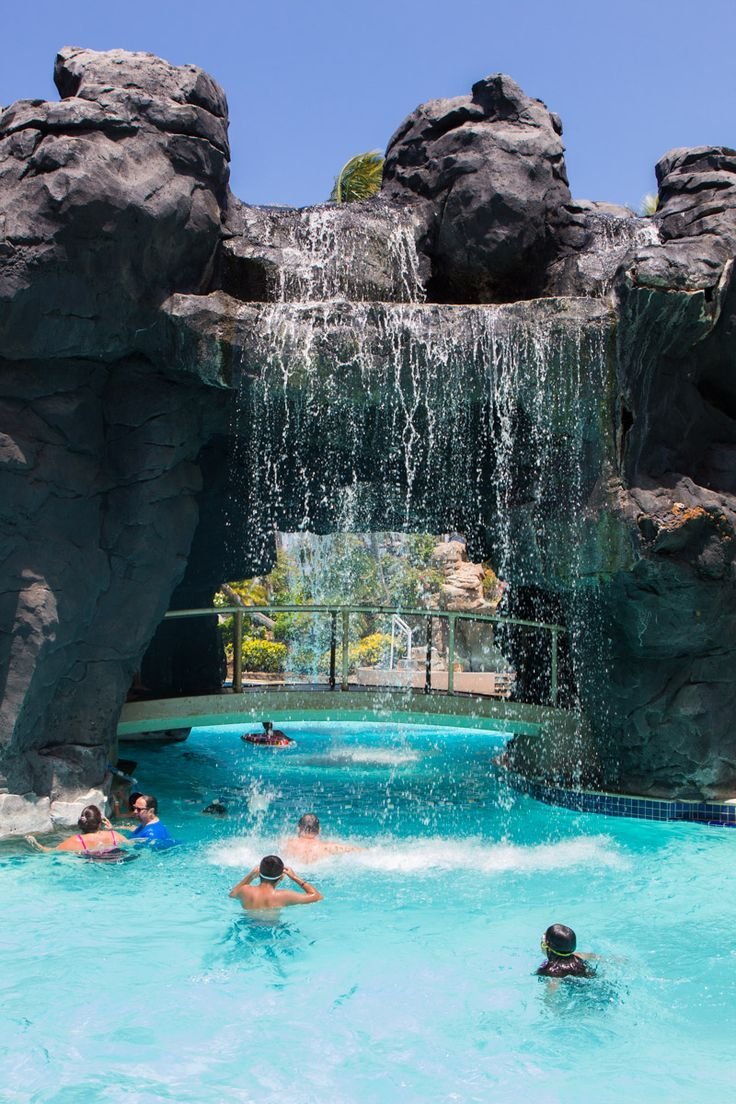 This may contain: several people are swimming in a large pool with water falling off the rocks and onlookers
