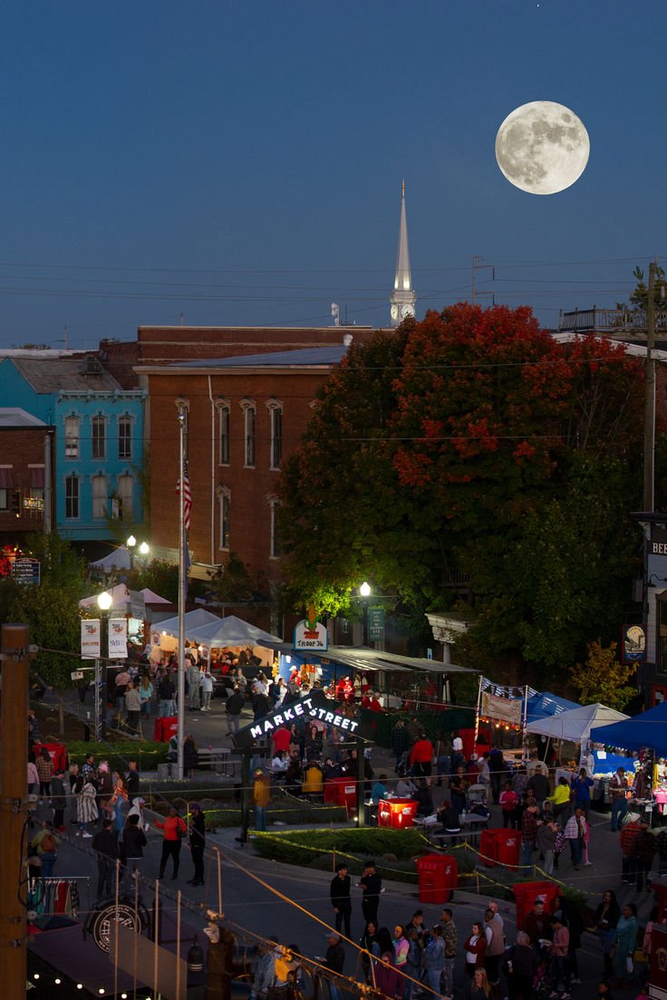 This may contain: the full moon shines brightly in the sky above an outdoor market with people walking around