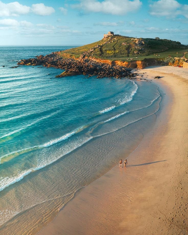 This may contain: two people are walking along the beach by the water's edge with an island in the background