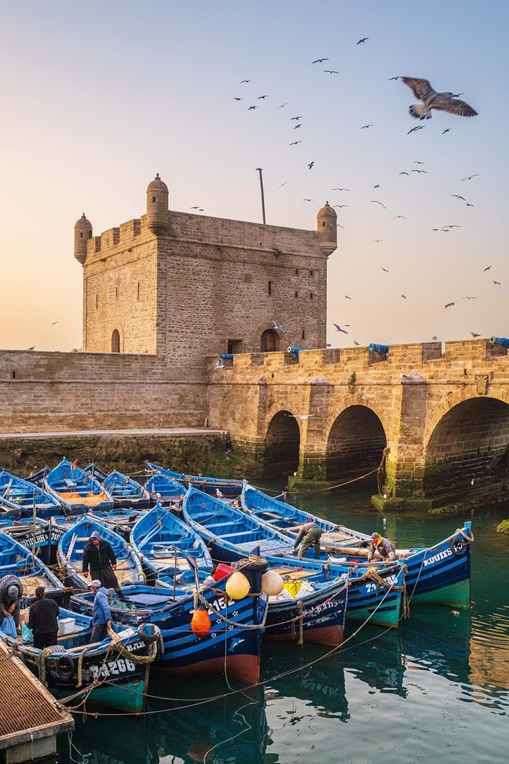 This may contain: there are many small boats docked in the water near a stone bridge and castle like structure
