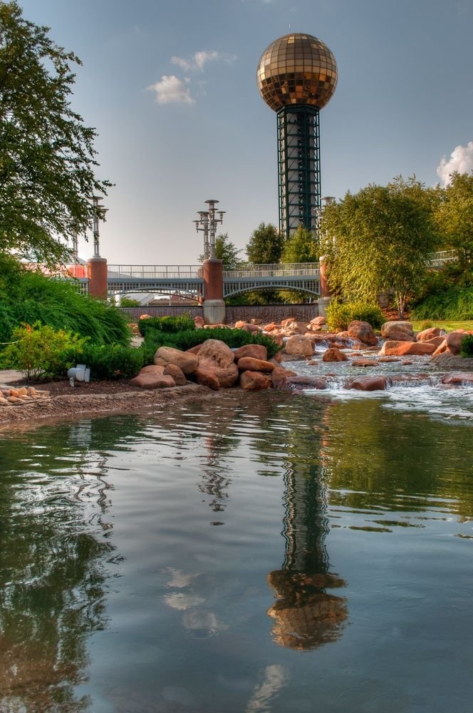 This may contain: the reflection of a tower in the water near rocks and trees, with a building in the background
