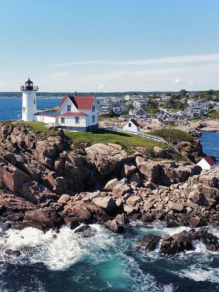 This may contain: a lighthouse on top of a rocky cliff next to the ocean with houses in the background
