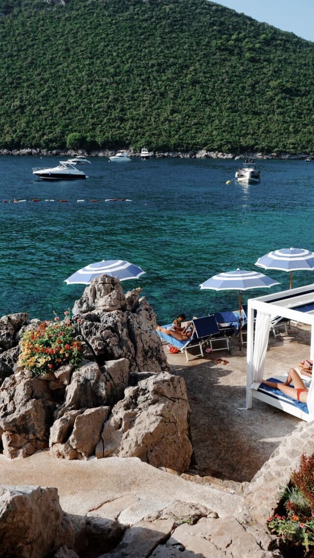 This may contain: people are lounging on the rocks by the water with umbrellas and boats in the background