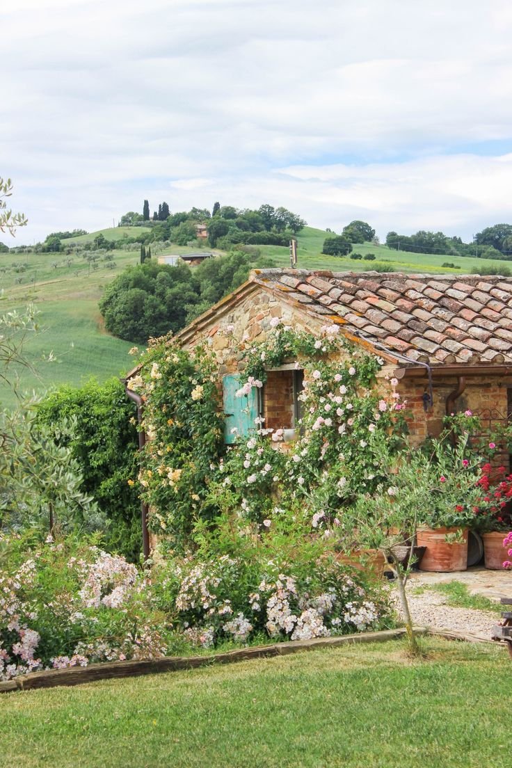 This may contain: an old stone house surrounded by flowers and greenery on a hill side with rolling hills in the background