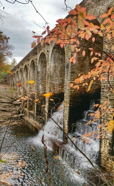 This may contain: an old stone bridge over a river with fall leaves on the trees and water running under it