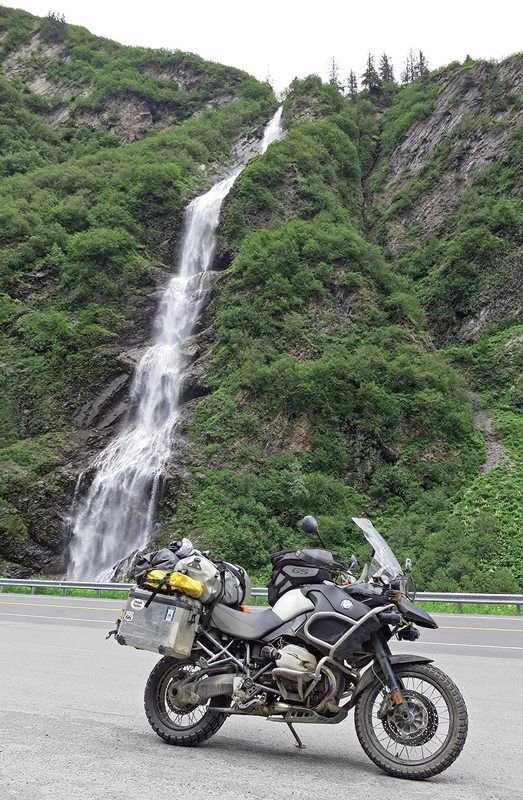 This may contain: a motorcycle parked next to a waterfall on the side of a road in front of a mountain