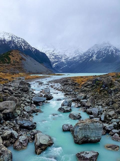 This may contain: a river running through a rocky valley surrounded by snow covered mountains and green grass, with rocks in the foreground