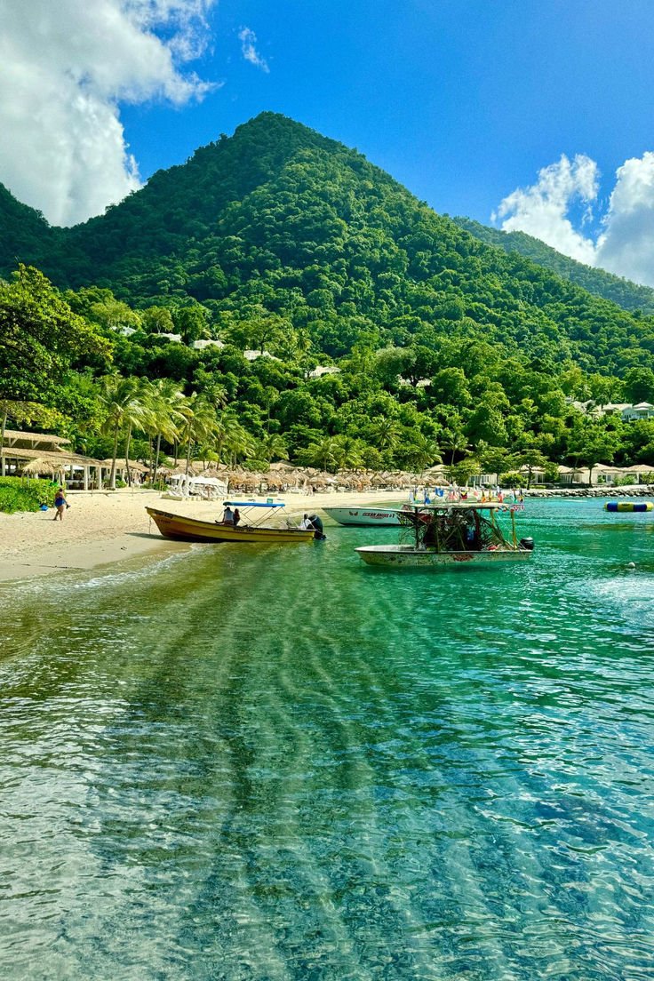 This contains: Scenic beach in St. Lucia with clear turquoise water, small boats anchored near the shore, and lush green mountains rising in the background under a bright blue sky with scattered clouds.