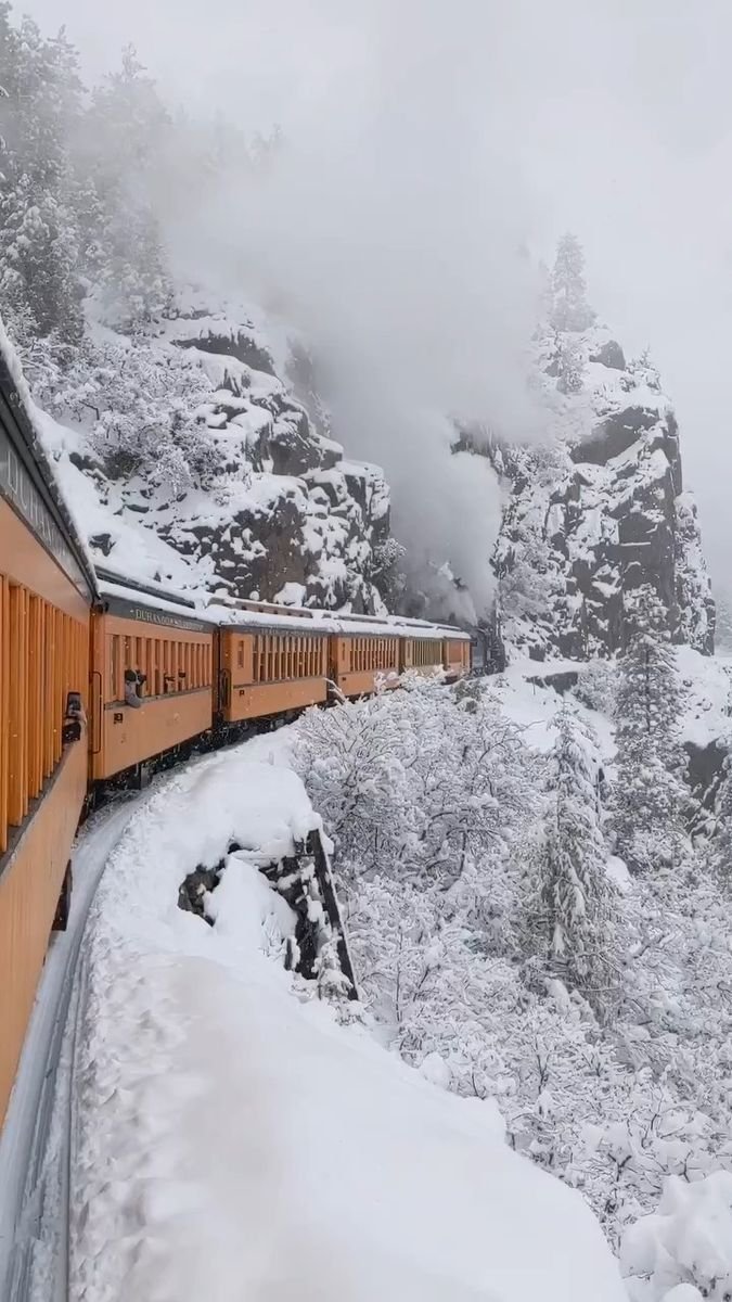 This may contain: a train traveling down tracks next to a snow covered mountain side under a cloudy sky