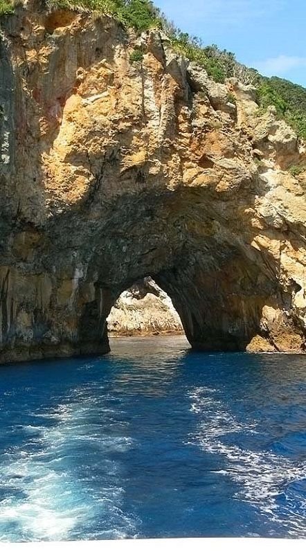 This may contain: an arch in the side of a cliff over looking blue water and land with trees on top