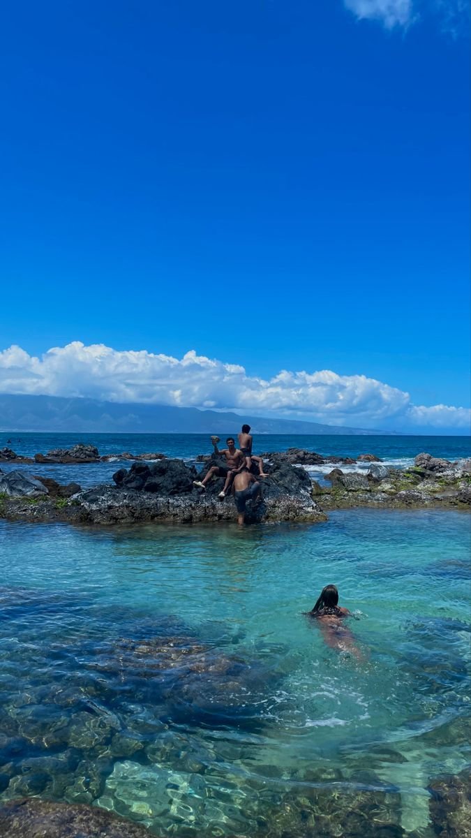 This may contain: two people sitting on rocks in the ocean with clear blue water and rocky shore behind them
