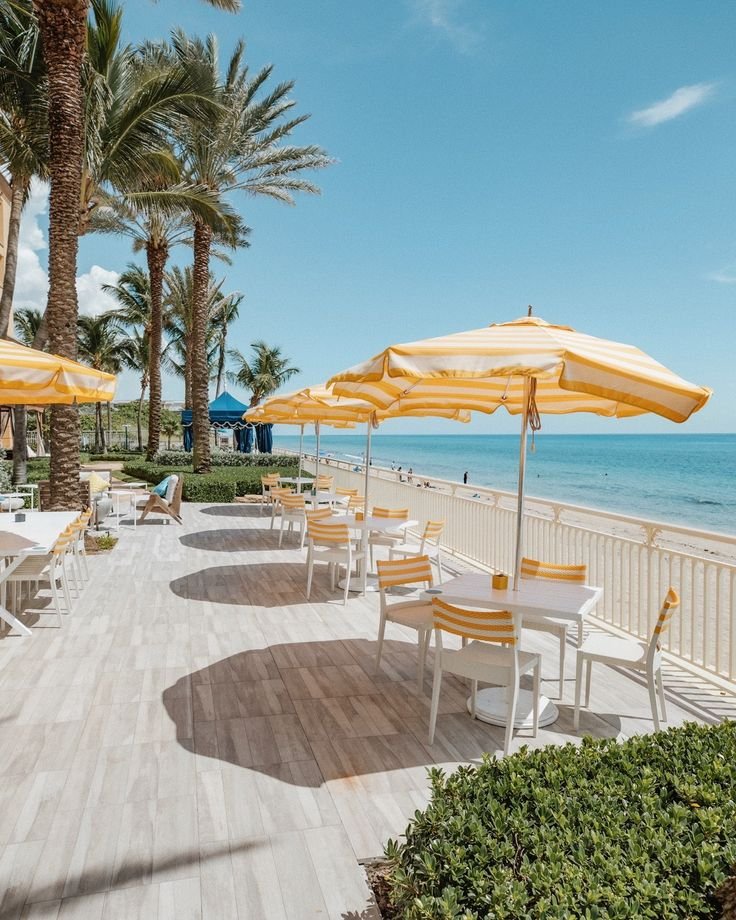 This may contain: tables and chairs with yellow umbrellas on the boardwalk by the ocean in front of palm trees