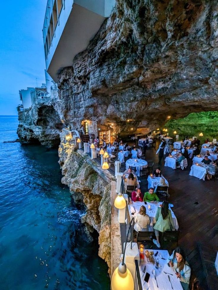 This may contain: an outdoor dining area next to the ocean with people sitting at tables and eating food
