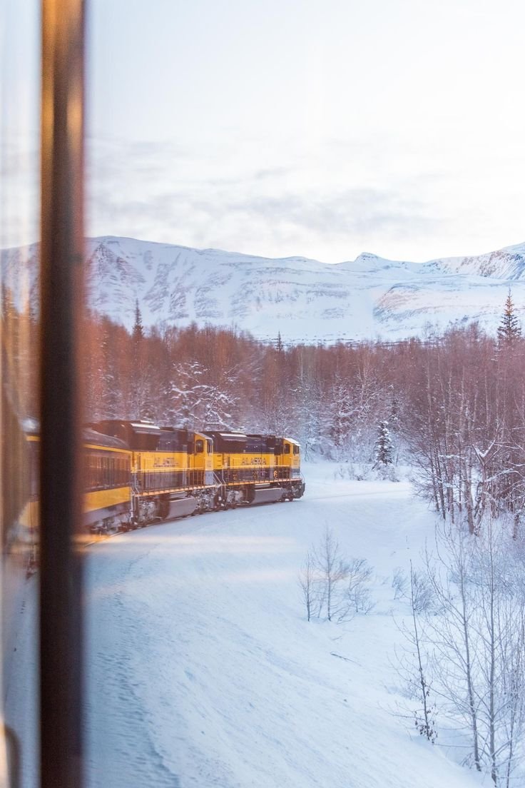 This may contain: a train traveling down tracks next to snow covered trees and mountains in the distance, seen through a window