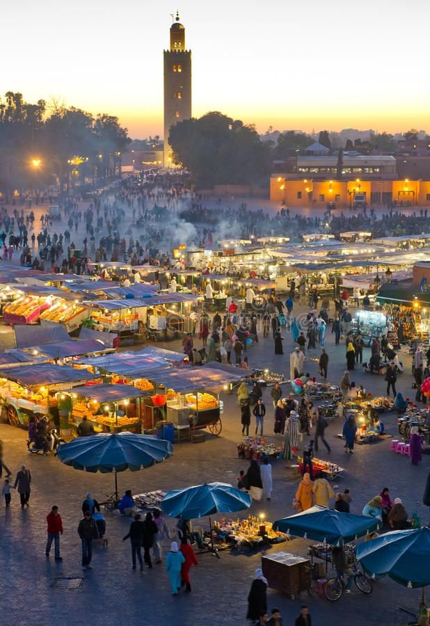 This may contain: an outdoor market with many stalls and people walking around it at sunset or sunrise time