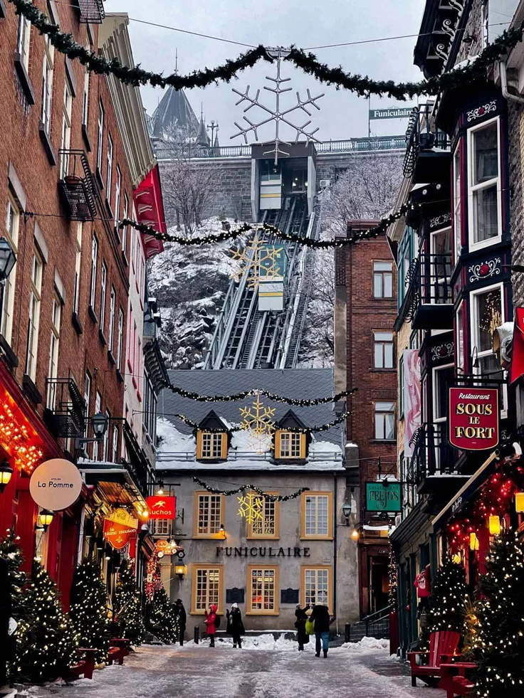 This may contain: people walk down a snowy street lined with buildings and decorated for the holiday season in an old european city