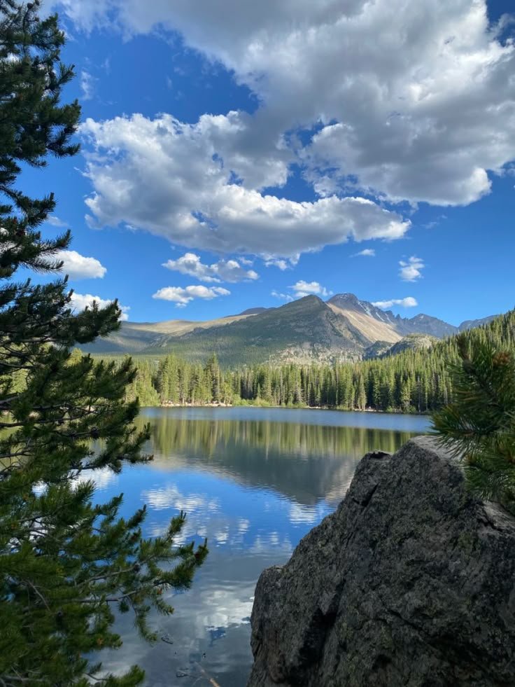 This may contain: a lake surrounded by trees and mountains under a cloudy blue sky with white fluffy clouds