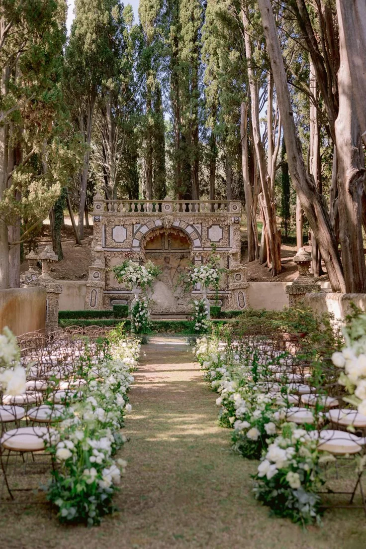 This may contain: an outdoor ceremony setup with white flowers and greenery on the aisle, surrounded by trees