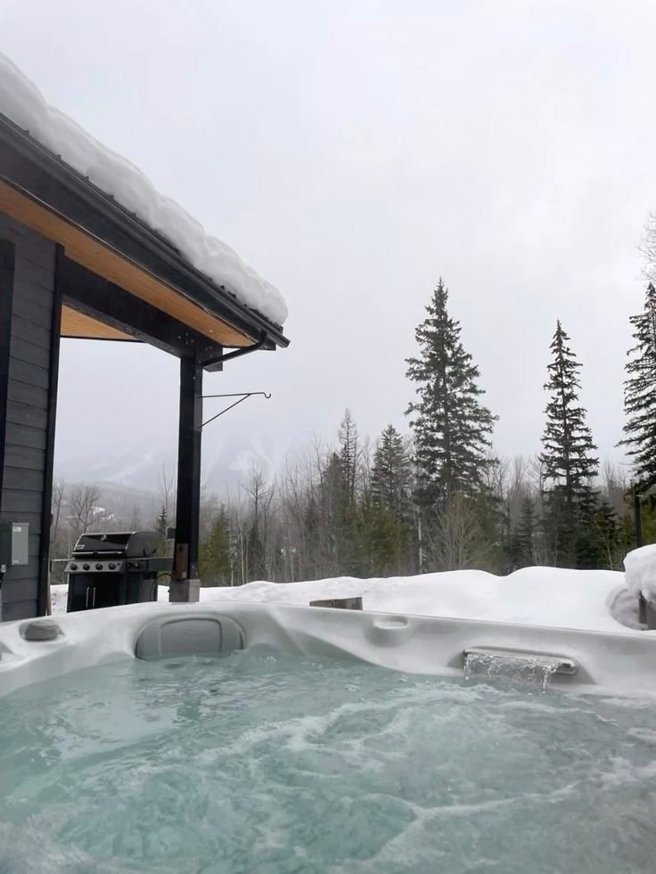 This may contain: an outdoor hot tub covered in snow next to a building with trees on the other side