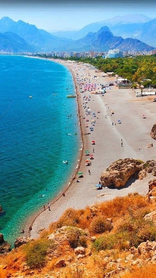 This may contain: the beach is crowded with people and mountains in the backgrouds, while blue water stretches into the distance