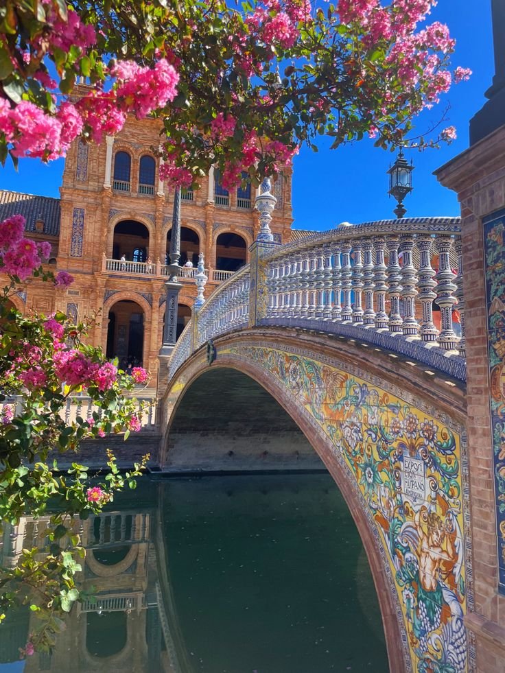 This may contain: an ornate bridge over a body of water with pink flowers in the foreground and a building on the other side