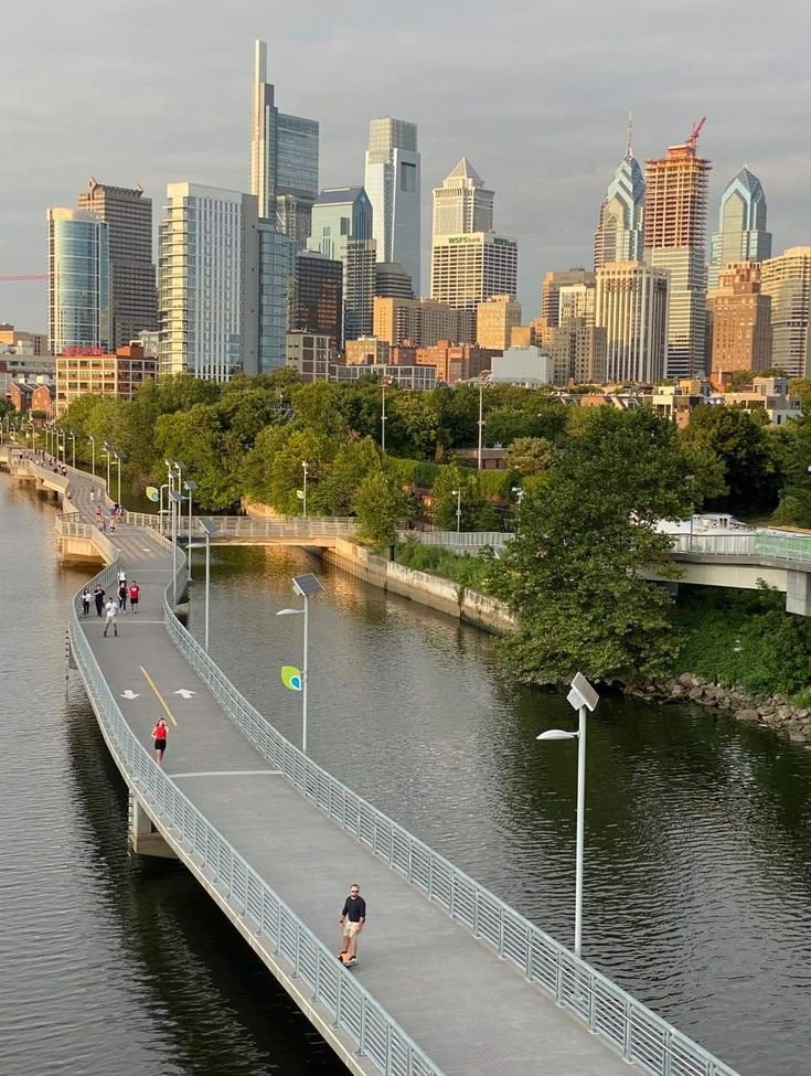 This may contain: people walking on a bridge over a river in front of a city skyline with tall buildings