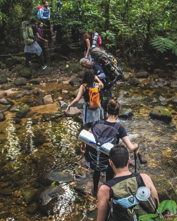 This may contain: a group of people walking across a stream in the woods with backpacks on their backs