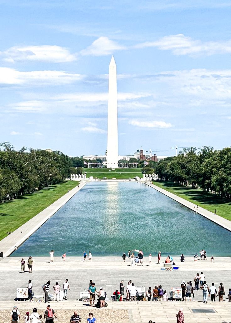 This may contain: people are standing in front of the reflecting pool at the washington monument, which is surrounded by trees and grass