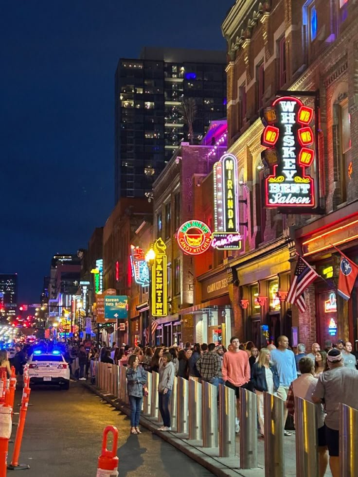 This may contain: a crowd of people standing on the side of a road next to tall buildings at night