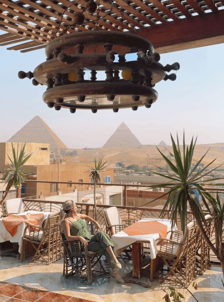 This may contain: a woman sitting at an outdoor table in front of the pyramids