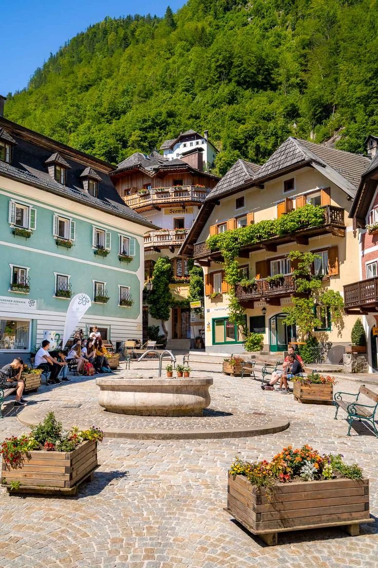 This may contain: people sitting on benches in the middle of an open courtyard with several buildings and green mountains behind them