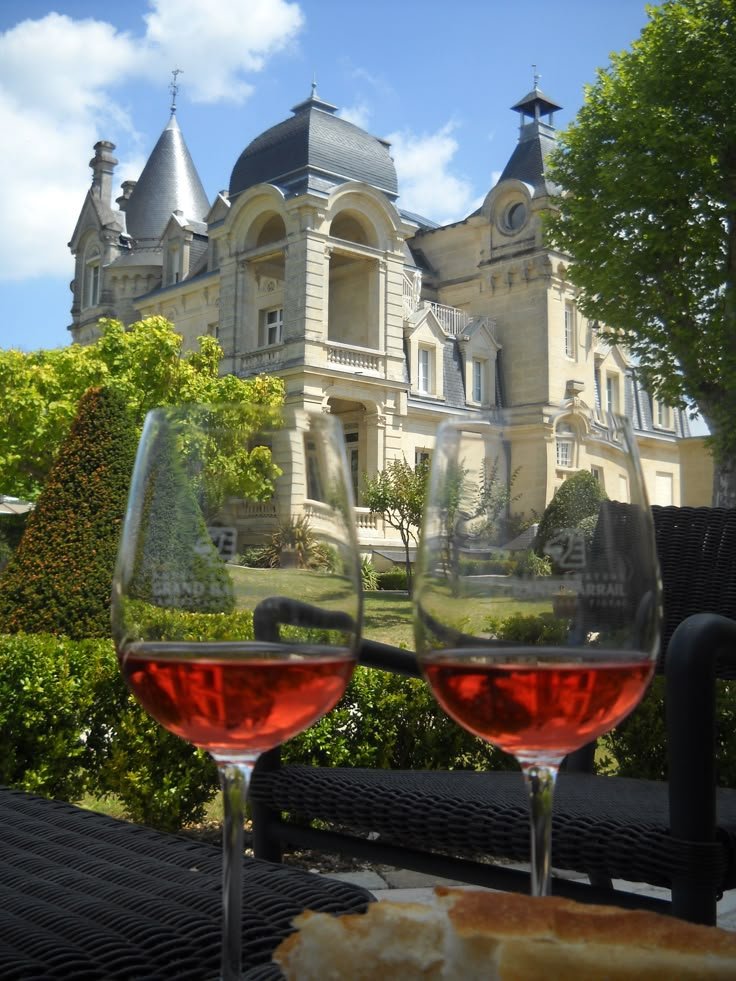 This may contain: two glasses of red wine sitting on top of a table in front of a large building