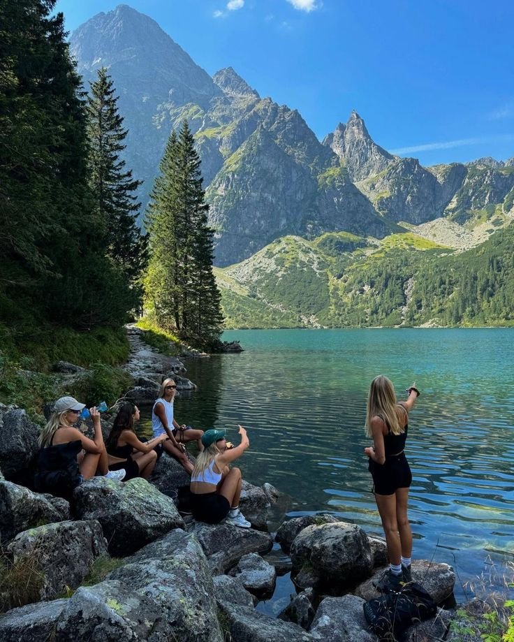 This may contain: some people are sitting on rocks by the water with mountains in the backgroud