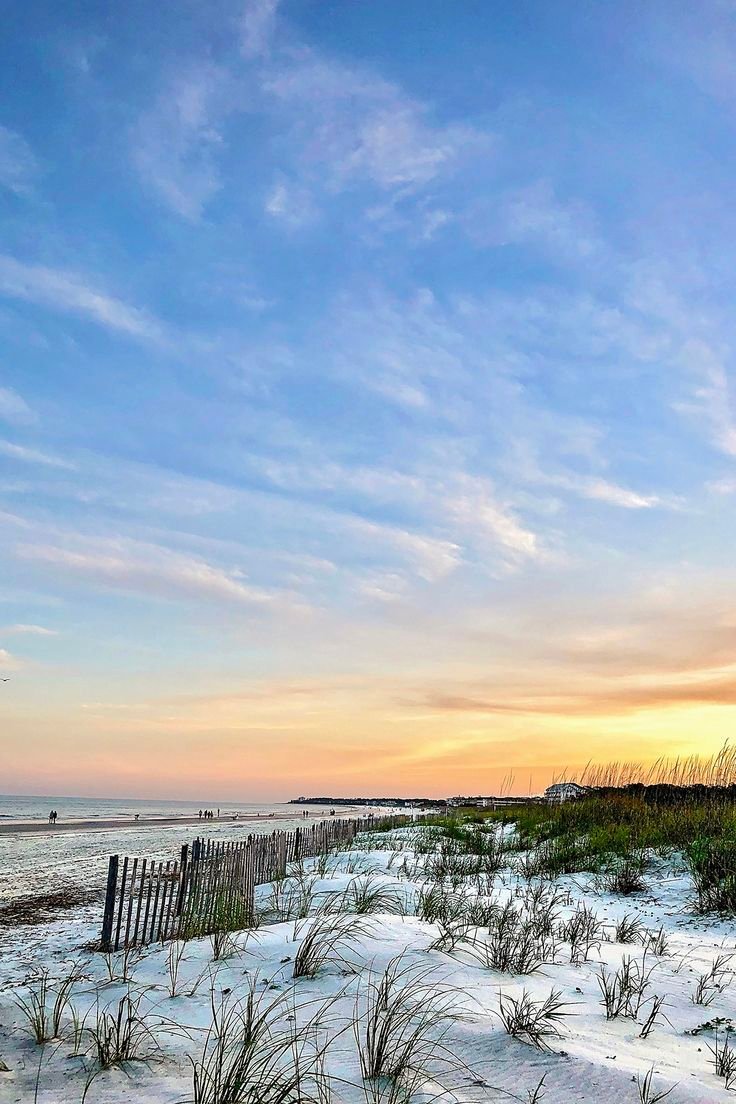 This contains: Scenic beach on Hilton Head Island with sand dunes illuminated by bright blue skies with trails of white clouds fading into a peach and orange sunset near the horizon line