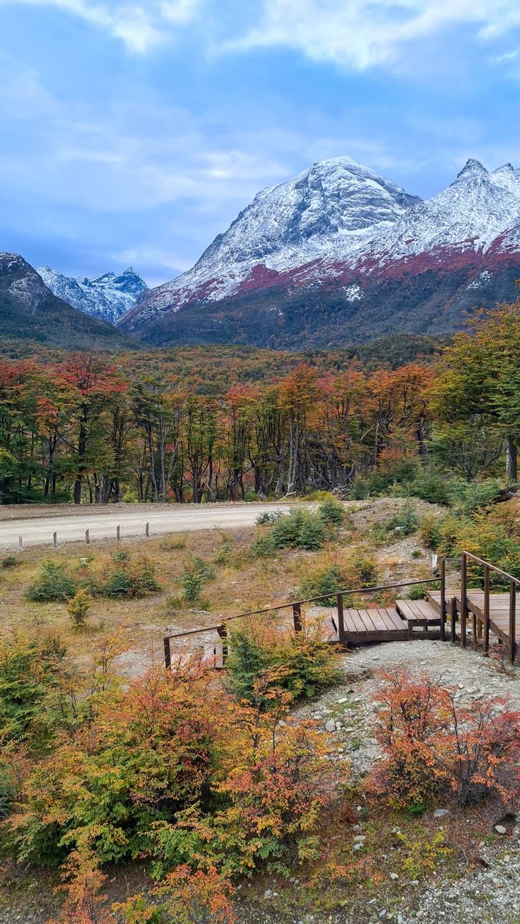 This may contain: the mountains are covered in snow and trees with autumn foliage on them, as well as a wooden bridge
