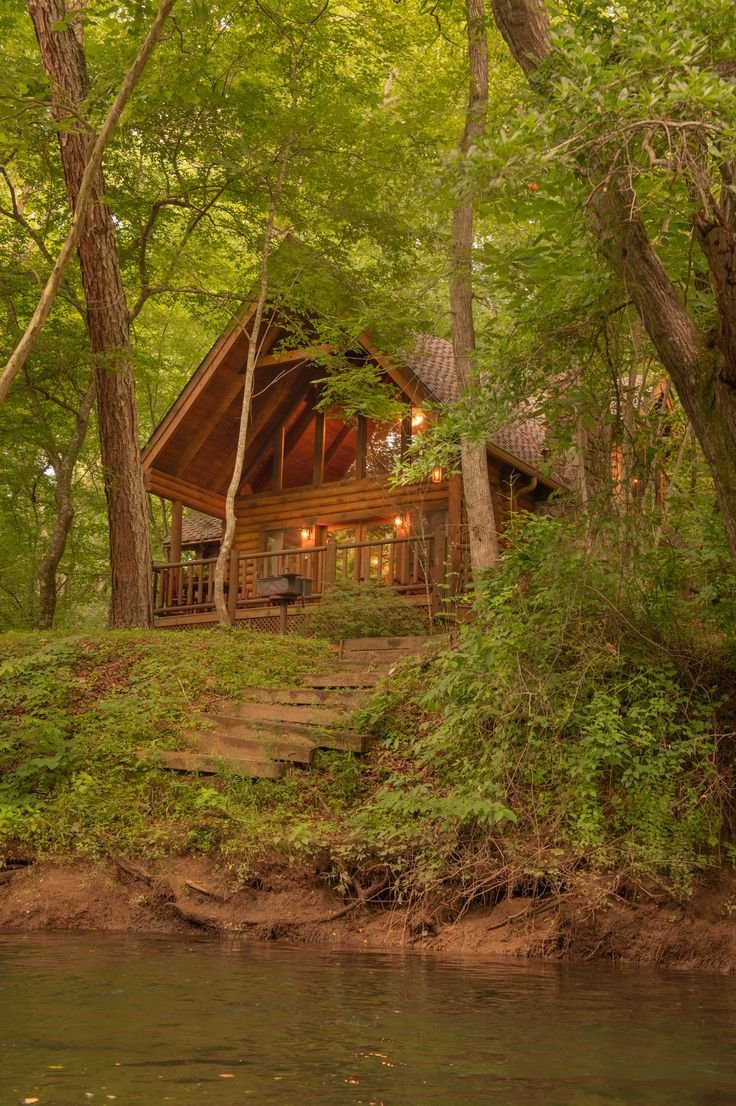 This may contain: a cabin in the woods next to a river with steps leading up to it and lights on