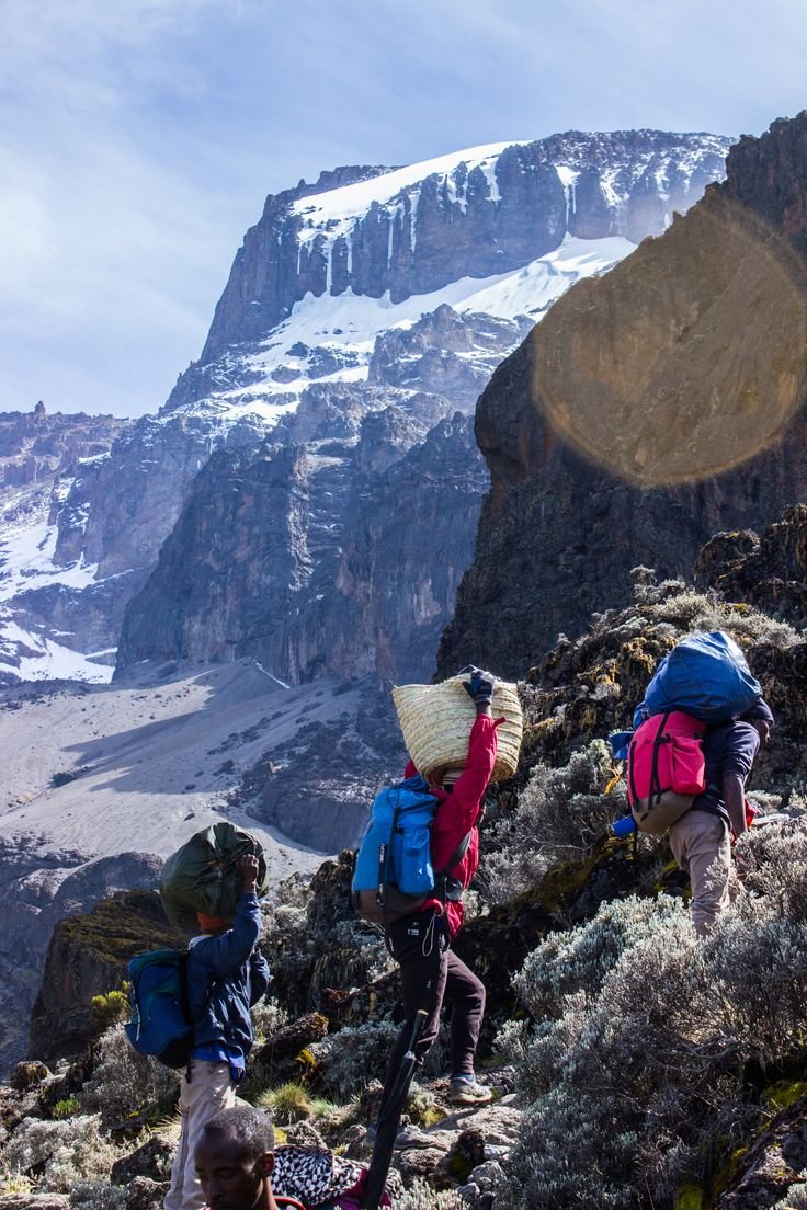 This may contain: three people with backpacks hiking up a mountain side in front of snow covered mountains