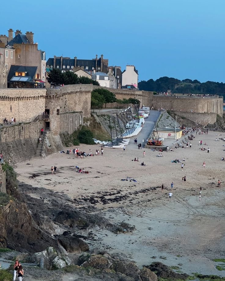 This may contain: many people are on the beach and in the water near some old buildings with towers