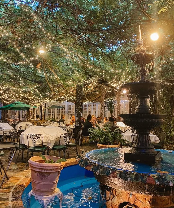 This may contain: an outdoor dining area with tables, chairs and a fountain in the center surrounded by string lights