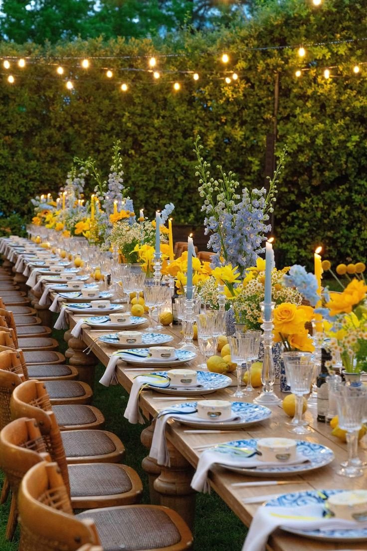 This may contain: a long table is set with yellow and white flowers in vases, candles, and plates